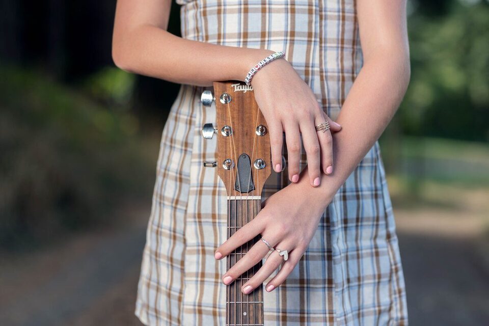 Close-up of Jette B's hands resting on the guitar headstock, wearing a beaded bracelet that reads Dear Yesterday.